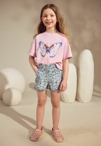 T-shirt rose clair avec un motif de papillon, associé à un short bleu clair à imprimé papillons et des sabots roses. L'arrière-plan présente des objets neutres et arrondis.