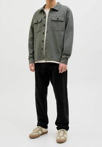 Oliwkowa koszula typu shirt jacket z guzikami, z dwiema kieszeniami na piersiach, w połączeniu z czarnymi spodniami i beżowymi sneakersami z białymi akcentami. Gładka tekstura.