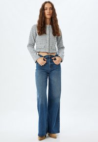 Grå texturerad kort jacka med knappar, kombinerad med högmidjade blå vida jeans. Modellen står med händerna på höfterna, neutral bakgrund.