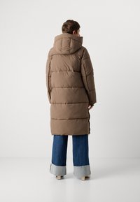 Manteau rembourré marron avec une capuche, design matelassé et fentes sur les côtés, associé à un jean en denim à jambe large avec des détails de poignets contrastants.