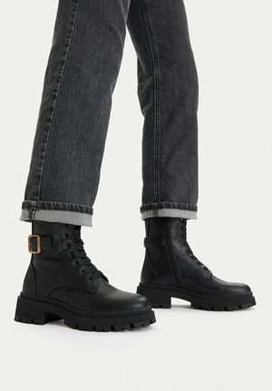 Schwarze Leder-Schnürstiefeletten mit seitlichen Schnallen, getragen zu aufgekrempelten dunkelgrauen Jeans vor einem einfarbigen hellen Hintergrund.