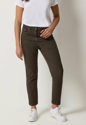 Nainen seisoo päällään tummanruskeat istuvat housut, valkoinen lyhythihainen paita ja valkoiset tennarit yksinkertaisen taustan edessä.