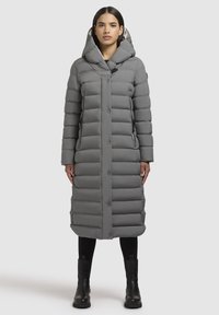 Grå puffjakke med hætte, med et langt, quiltet design og trykknapper. Båret over sorte leggings og ankelstøvler. Glat tekstur.