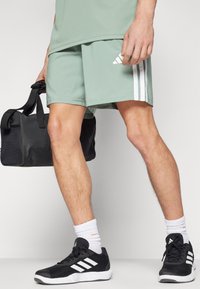 Hellgrüne Sportshorts mit weißen Seitenstreifen, kombiniert mit schwarzen Turnschuhen und einer schwarzen Sporttasche. Weiße Sneakersocken vervollständigen den Look.