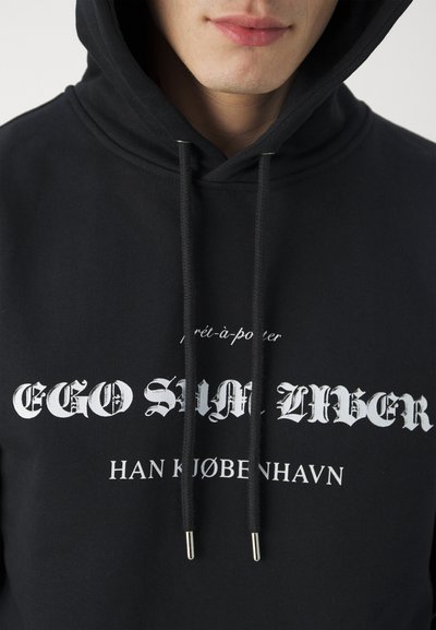 Hoodie noir avec texte blanc, cordons de serrage avec embouts en métal, et une texture douce. Le texte comprend "EGO SUM LIBER" et des détails de la marque.