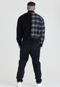 Homme avec un tatouage au cou portant une veste à carreaux noire et blanc, un jean noir et des baskets blanches, debout de dos sur un fond uni.