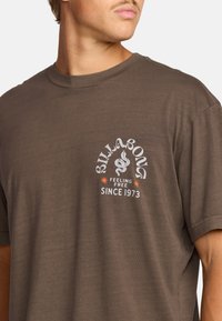 Ruda medvilninė marškinėliai su trumpomis rankovėmis, su balta grafika, gyvatės motyvas ir tekstu: "BILLABONG FEELING FREE SINCE 1973."