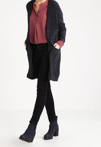 Persona che indossa un lungo cardigan grigio scuro, un top bordeaux con bottoni, pantaloni skinny neri e stivaletti con tacco blu scuro, in piedi con le mani nelle tasche.