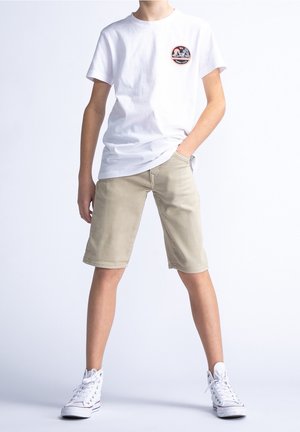 T-shirt en coton blanc avec un logo circulaire sur la poitrine, associé à un short beige clair. Le mannequin porte des baskets blanches. Design simple et coupe droite.