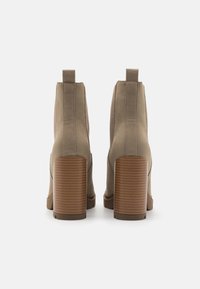 Une paire de bottes cheville en daim taupe avec des talons blocs empilés et des languettes, montrée de dos sur un fond blanc.