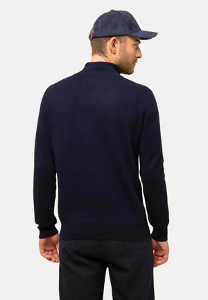 Marineblauer Strickpullover mit gerippter Textur, ausgestattet mit einem klassischen Kragen und langen Ärmeln. Kombiniert mit dunklen Hosen und einer passenden Mütze.