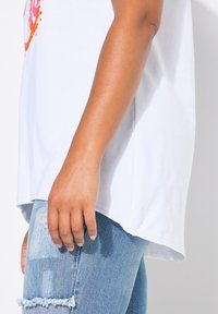 T-shirt en coton blanc avec fentes latérales, présentant un imprimé abstrait coloré, associé à un jean bleu clair déchiré.