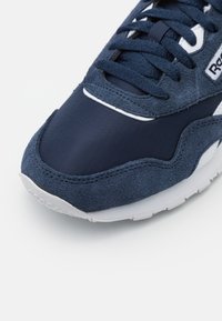 Navy blue sneaker featuring a combination of suede and textile materials, with a white rubber sole and textured accents. Lace-up design.