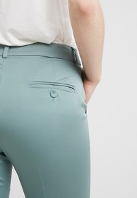 Pantalones ajustados en color azul verdoso con una textura suave, que cuentan con un bolsillo trasero con detalle de botón y una trabilla en la cintura.