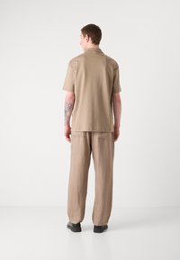 Beige polo shirt met korte mouwen, gecombineerd met lichtbeige broek, gedragen met zwarte schoenen. Glad textuur, losse pasvorm, zichtbare naaddetail aan de achterkant.