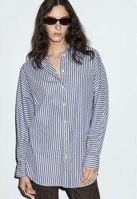 Camicia a righe con strisce verticali bianche e navy, maniche lunghe, vestibilità comoda e colletto. Occhiali da sole marroni e tessuto testurizzato.