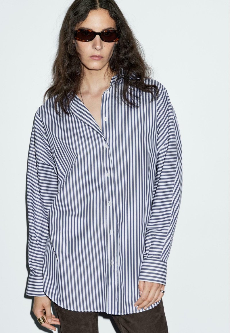 Camicia a righe con strisce verticali bianche e navy, maniche lunghe, vestibilità comoda e colletto. Occhiali da sole marroni e tessuto testurizzato.