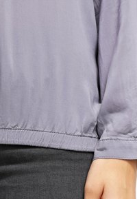 Blouse grise, légère, à manches longues, avec des poignets froncés et une texture douce. Ourlet fini avec une bande élastique pour un look cintré.