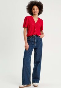 Chemisier rouge à boutons avec des manches courtes bouffantes et des détails en tissu à œillets, associé à un jean denim large taille haute bleu et des chaussures légères.