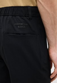 Κοντινό πλάνο στην τσέπη μαύρου παντελονιού με το λογότυπο "Porsche BOSS" ανάγλυφο πάνω από την τσέπη, στο ισχίο ενός ατόμου με μερικώς ορατό μπράτσο.