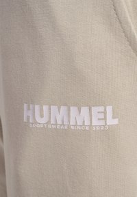 Beige Sportbekleidung mit strukturiertem Stoff. Der Markenname „HUMMEL“ ist in fettgedruckten, weißen Buchstaben dargestellt, darunter steht „SPORTBEKLEIDUNG SEIT 1923“. 