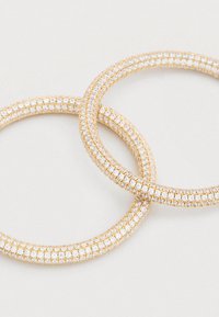 Náušnice v zlatej farbe v tvare kruhu, zdobené kryštálom pripomínajúcim diamant, so štruktúrovaným povrchom a kruhovým dizajnom s prekrývajúcimi sa prvkami.