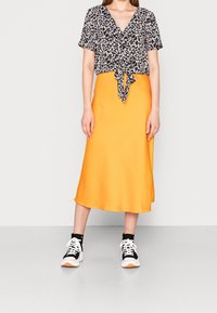 Frau trägt eine schwarze kurzärmelige Bluse mit Blumendruck und Krawattenfront, einen leuchtend orangefarbenen Midirock, schwarz-weiße Sneaker und steht auf einem grauen Boden.