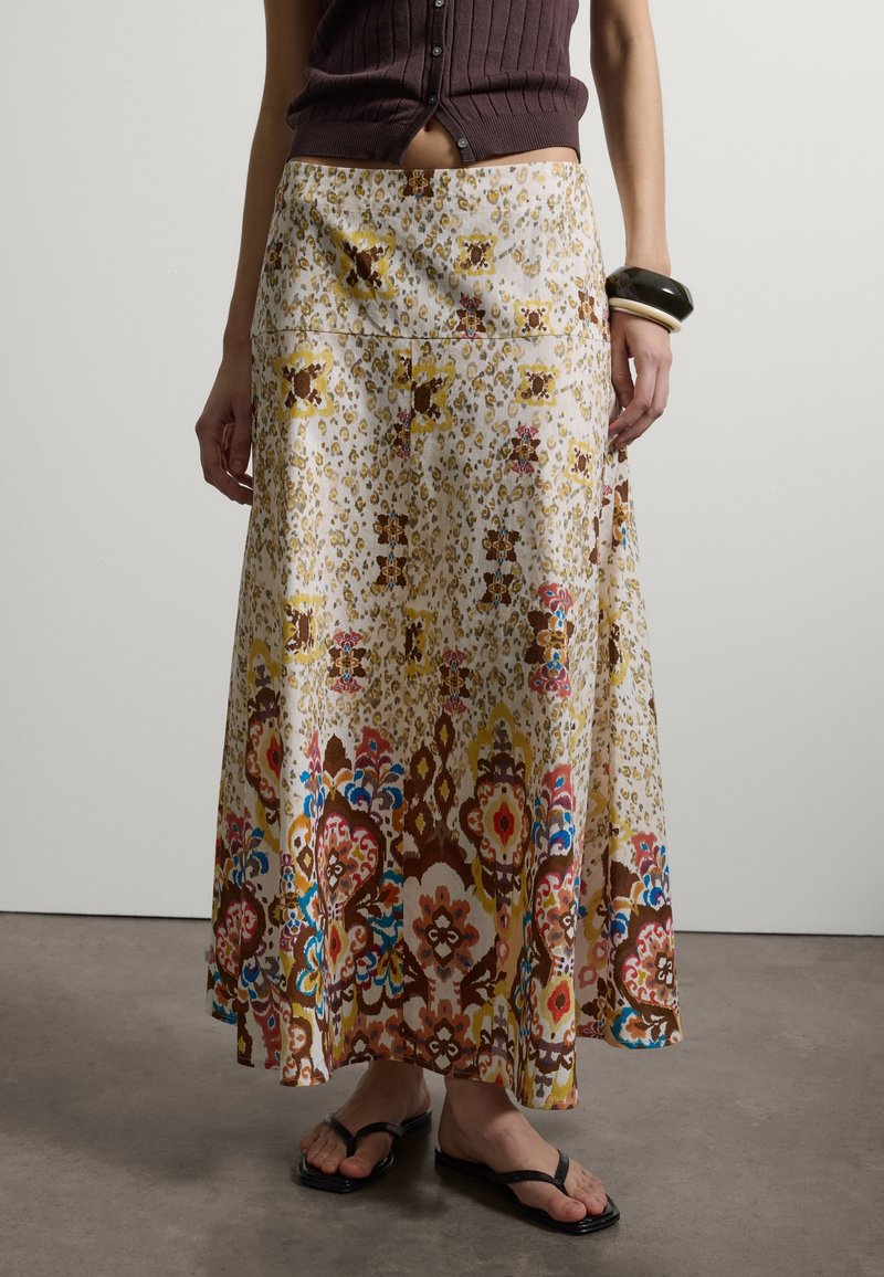 Frau trägt einen langen, gemusterten Rock mit Blumen- und geometrischen Mustern, ein braunes ärmelloses Oberteil, Armreifen an einem Handgelenk und schwarze Sandalen.