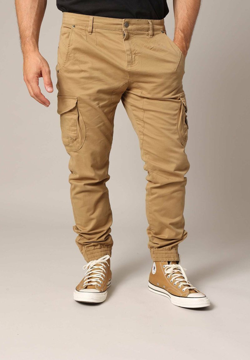Khaki cargobroek met taps toelopende boorden, voorzien van meerdere zakken en een gladde textuur. Gecombineerd met lichtbruine hoge sneakers.