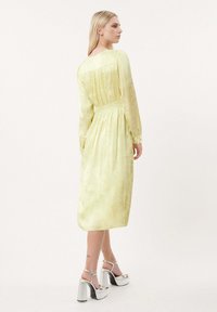 Gelbes, langärmeliges Midikleid mit gerafftem Bund, strukturierter Stoff und subtiler Glanz. Silberne Plateauschuhe sind unten sichtbar.