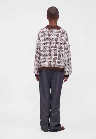Personne aux cheveux courts portant un pull marron et blanc à motifs, un pantalon large sombre et des chaussures marron, debout de dos sur un fond uni.