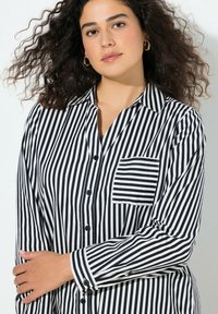 Camicia a maniche lunghe con bottoni, a righe bianche e nere, con una tasca sul petto e una texture del tessuto morbida. Il motivo presenta righe verticali.