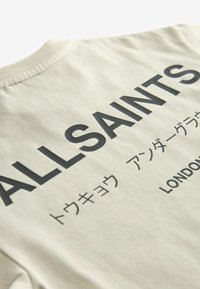 Beige puuvillane T-särk, millel on suur must tekst "ALLSAINTS" ja lisatekst jaapanikeeles. Sile tekstuur ja ümar kaelusdisain.