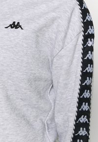 Sudadera gris claro con el logotipo de Kappa en blanco y negro en el pecho y una franja repetida con el logotipo de Kappa que recorre la manga.