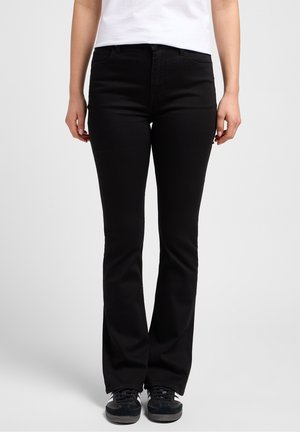 Vrouw met zwarte bootcut jeans, zwart-witte sneakers en een wit topje, staand tegen een effen lichte achtergrond.
