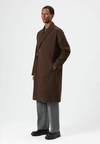 Bruine wollen overjas met een reverskraag, enkele knoop, zijzakken en een metalen accent, gecombineerd met grijze broek en zwarte schoenen.