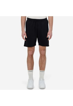 Homme portant un short noir à taille élastique, des chaussettes blanches montantes avec texte noir, et des baskets blanches, debout devant un fond blanc uni.