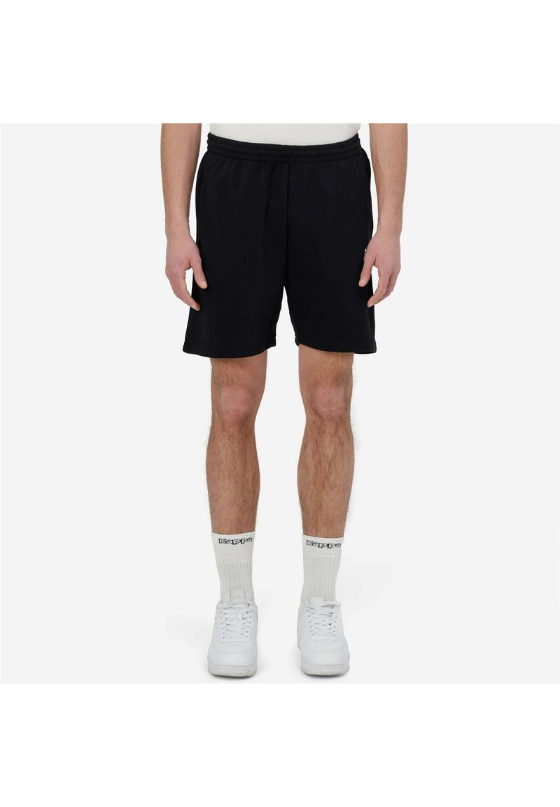 Homme portant un short noir à taille élastique, des chaussettes blanches montantes avec texte noir, et des baskets blanches, debout devant un fond blanc uni.