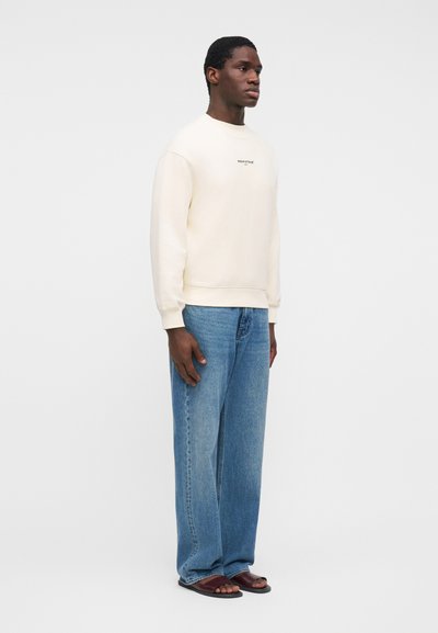 Άνδρας που στέκεται φορώντας μια κρεμ φούτερ, φαρδιά μπλε τζιν και καφέ σανδάλια σε απλό λευκό φόντο.