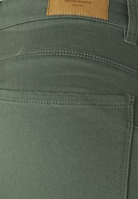 Jeans de denim verde con una textura suave, costuras prominentes y una etiqueta de cuero con la marca "VERO MODA" cerca de la cintura.