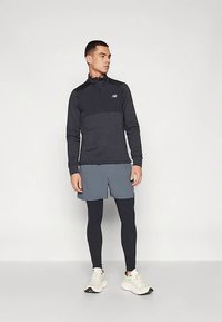 Juodas ir pilkas ilgomis rankovėmis užsegamas džemperis, mėlyni šortai, juodos tamprės ir balti sportbačiai; ant krūtinės - New Balance logotipas.