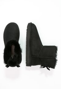 Czarne zamszowe botki za kostkę z futrzanym wyściółką, płaską gumową podeszwą i dekoracyjnymi kokardkami z tyłu. Okrągły czubek i miękka tekstura.