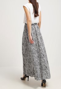 Kvinna som bär en ärmlös vit blus, långkjol med svart-vitt mönster och svarta högklackade skor, står med ryggen vänd framåt.