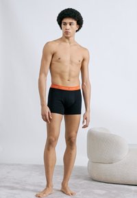 Μαύρα μπόξερ με πορτοκαλί μέση, με άνετη εφαρμογή και απαλό υλικό. Το μοντέλο στέκεται σε ανοιχτό γκρι χαλί.