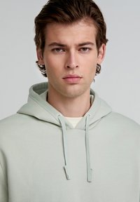 Lichtgroene hoodie met een voorzak in kangoeroestijl, een capuchon met trekkoord en een zachte textuur. Gedragen over een crèmekleurig shirt, minimalistisch ontwerp.