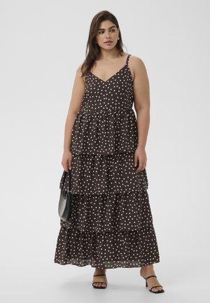 Naine, kellel on seljas musta värvi varrukateta maxi-kleit valgete täppidega, hoides käes musta käekotti ja kandes musta kontsaga sandale.