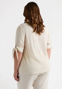 Helles, cremefarbenes Blusenshirt aus strukturiertem Stoff, mit Dreiviertelärmeln und gebundenem Detail an den Ärmeln sowie einem lässigen Schnitt.