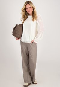 Transparante witte blouse met bloembroderie en elastische manchetten, gecombineerd met beige geruite wijde broek en een bruine leren tas.