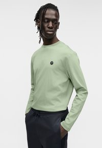 Μακρυμάνικο, ανοιχτό πράσινο μπλουζάκι με στρογγυλή λαιμόκοψη και μαύρο λογότυπο στο στήθος, συνδυασμένο με σκούρο παντελόνι με ελαστική μέση.