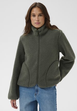 Nő zöld, cipzáras polárdzsekiben, magas gallérral és kék farmernadrágban, egyszínű világos háttér előtt áll.